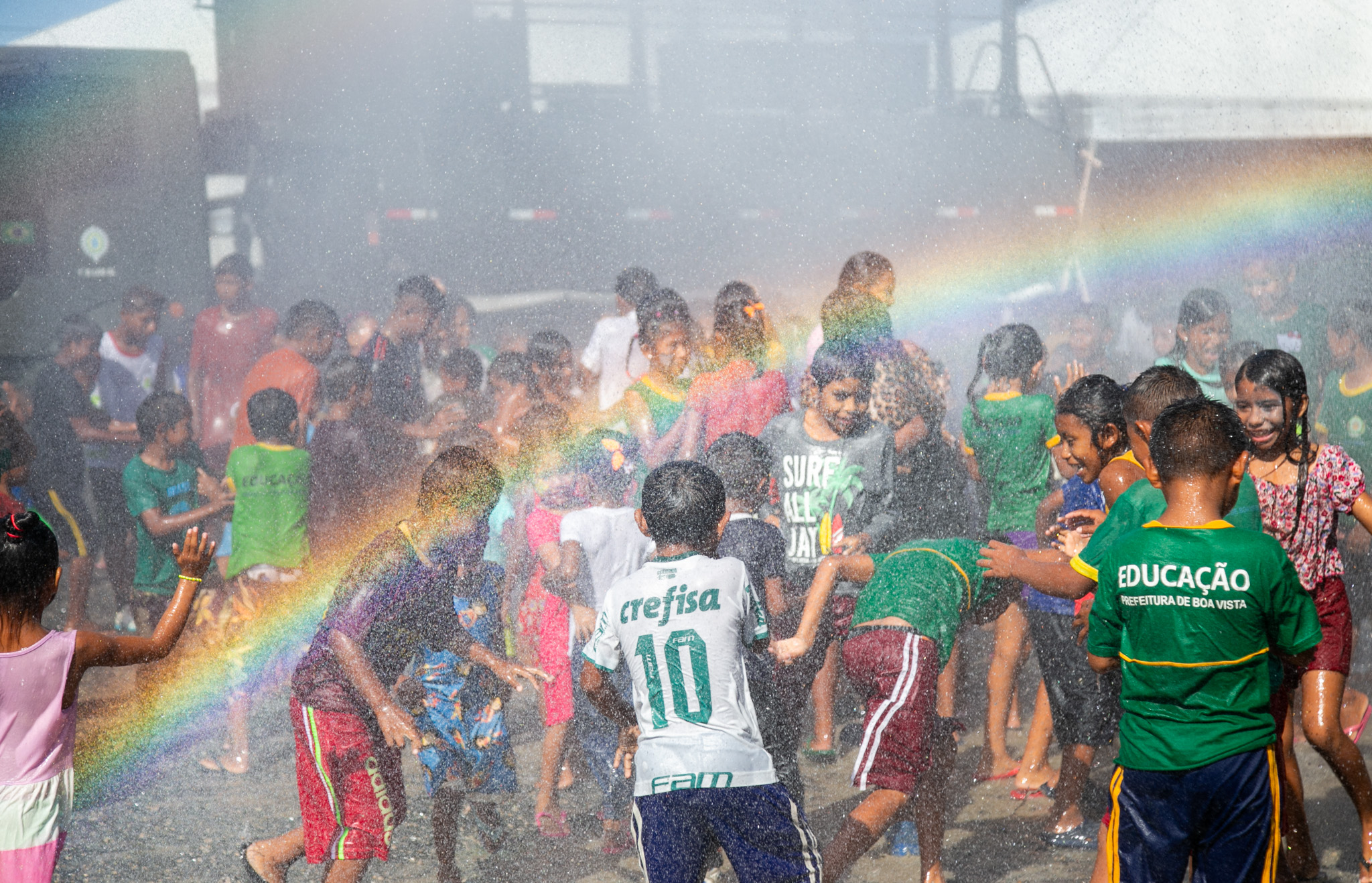 Children playing under a water spray with a visible rainbow, smiling and enjoying a sunny day.