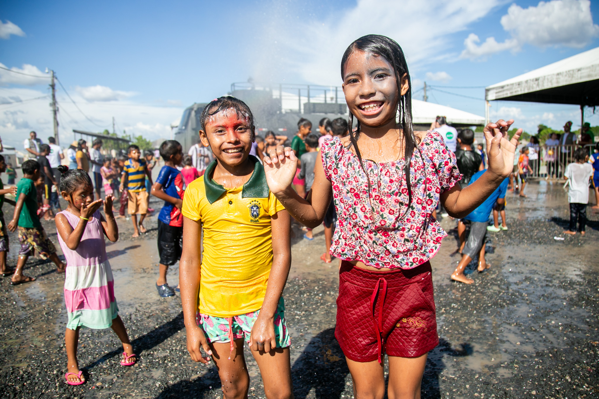 Children enjoying a water fight outdoors, smiling, and wearing colorful clothes on a sunny day, with others playing in the background.