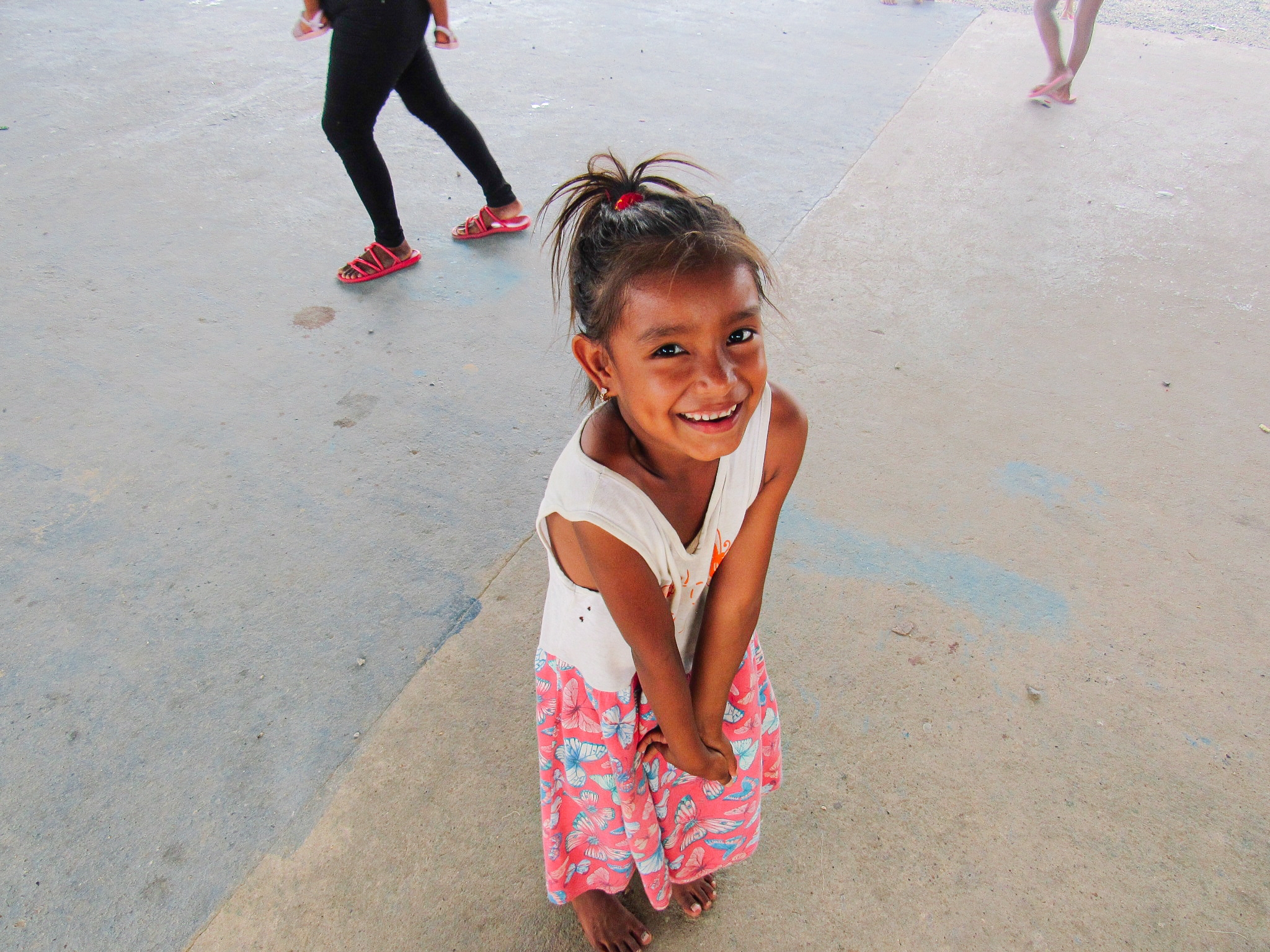 Smiling young girl with a ponytail, wearing a white top and colorful skirt, stands barefoot on a concrete floor. People walk in the background.