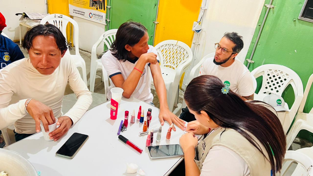 Four people at a table, one applying nail polish while others observe. Various nail polish bottles are scattered on the table.
