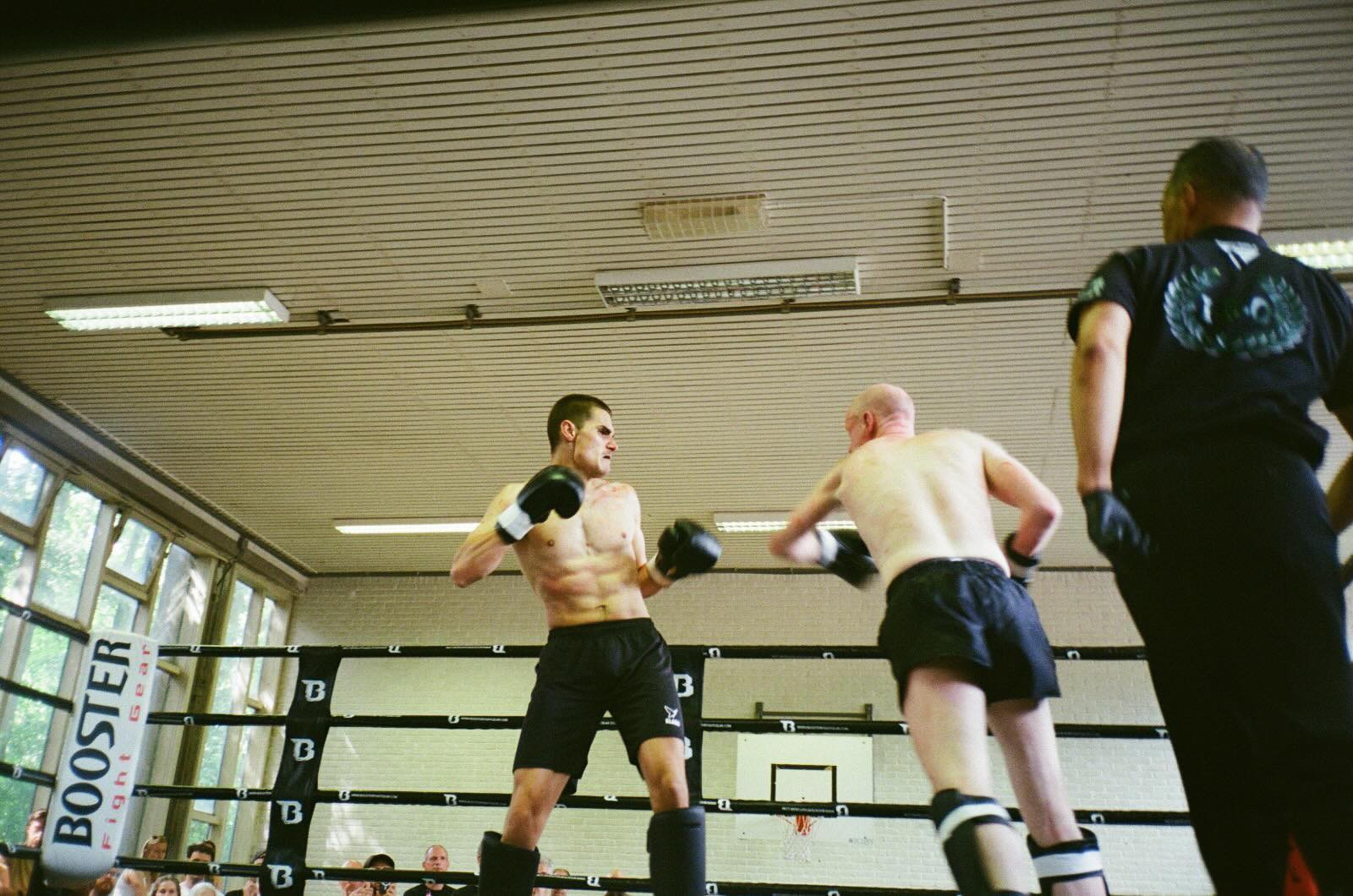 Two boxers spar in a ring, overseen by a referee in a gym with high windows and spectators visible in the background.