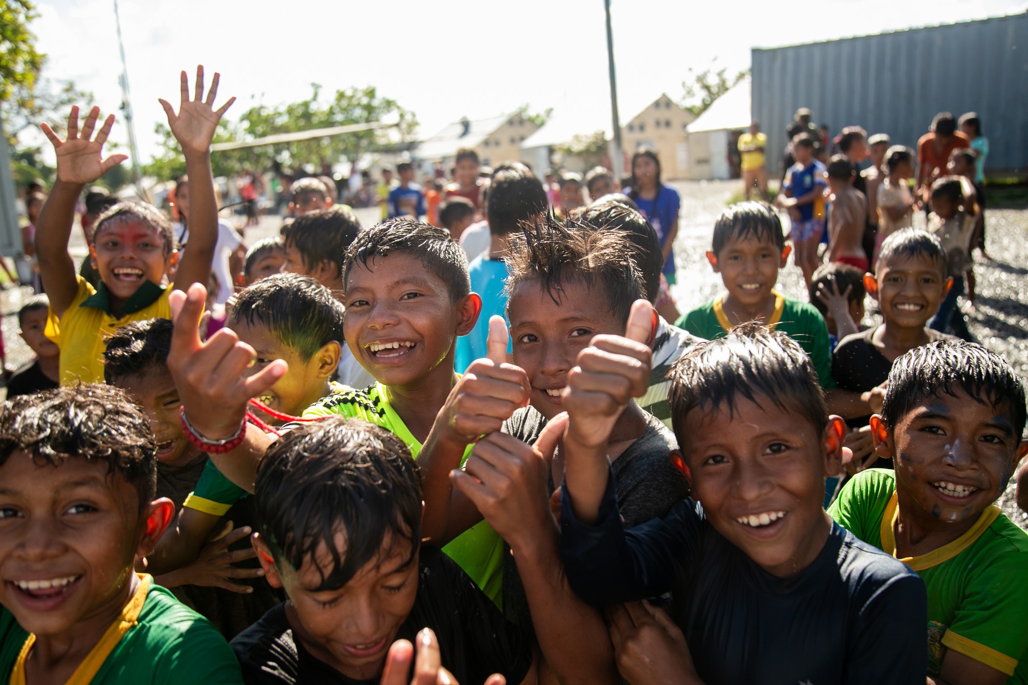 Group of joyful children outdoors, smiling and giving thumbs up, with some raising their hands in the air, dressed in colorful shirts.