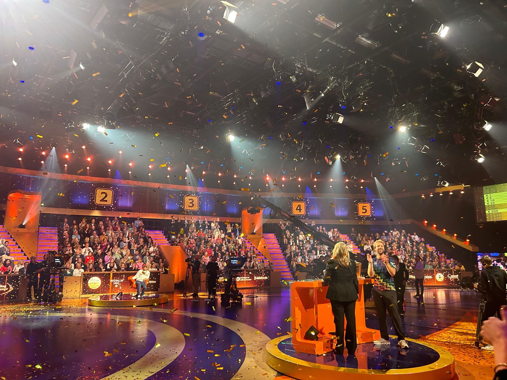 A vibrant TV game show set with hosts on stage, audience in stands, and colorful confetti falling from above.