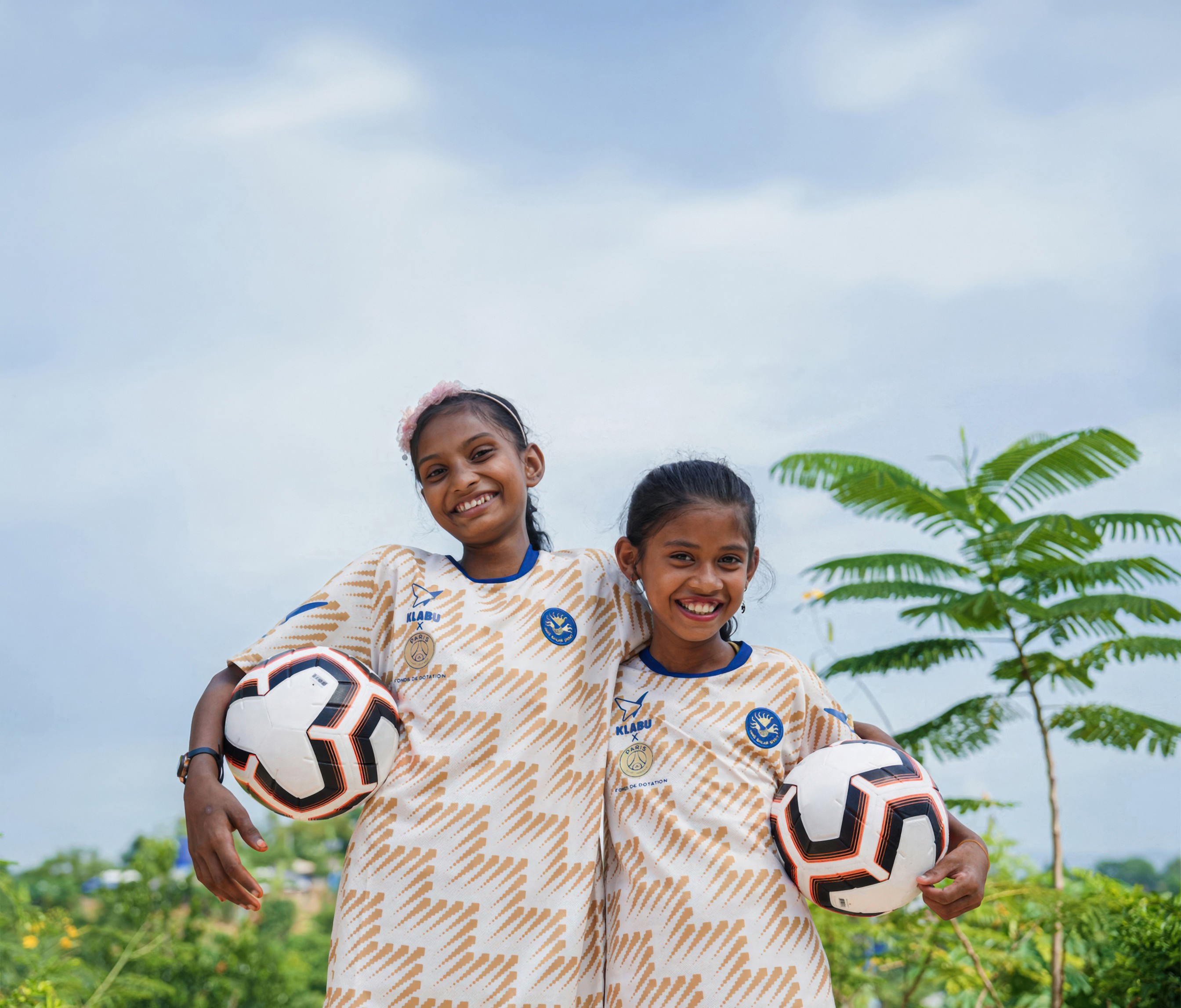 Two smiling girls in matching jerseys, holding soccer balls, stand outdoors against a blue sky and greenery.