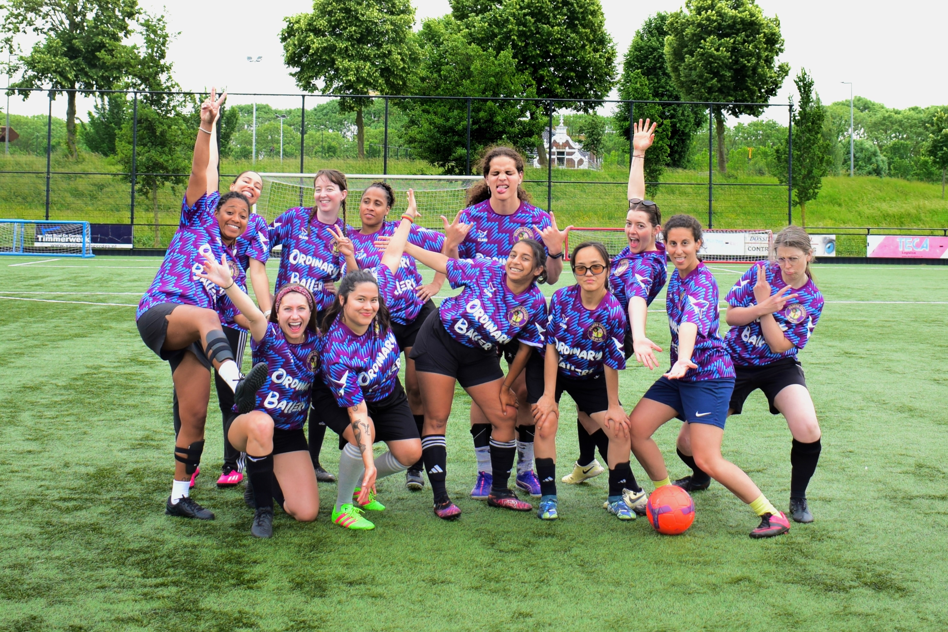 A group of smiling women soccer players in colorful jerseys pose energetically on a grassy field with a ball nearby.