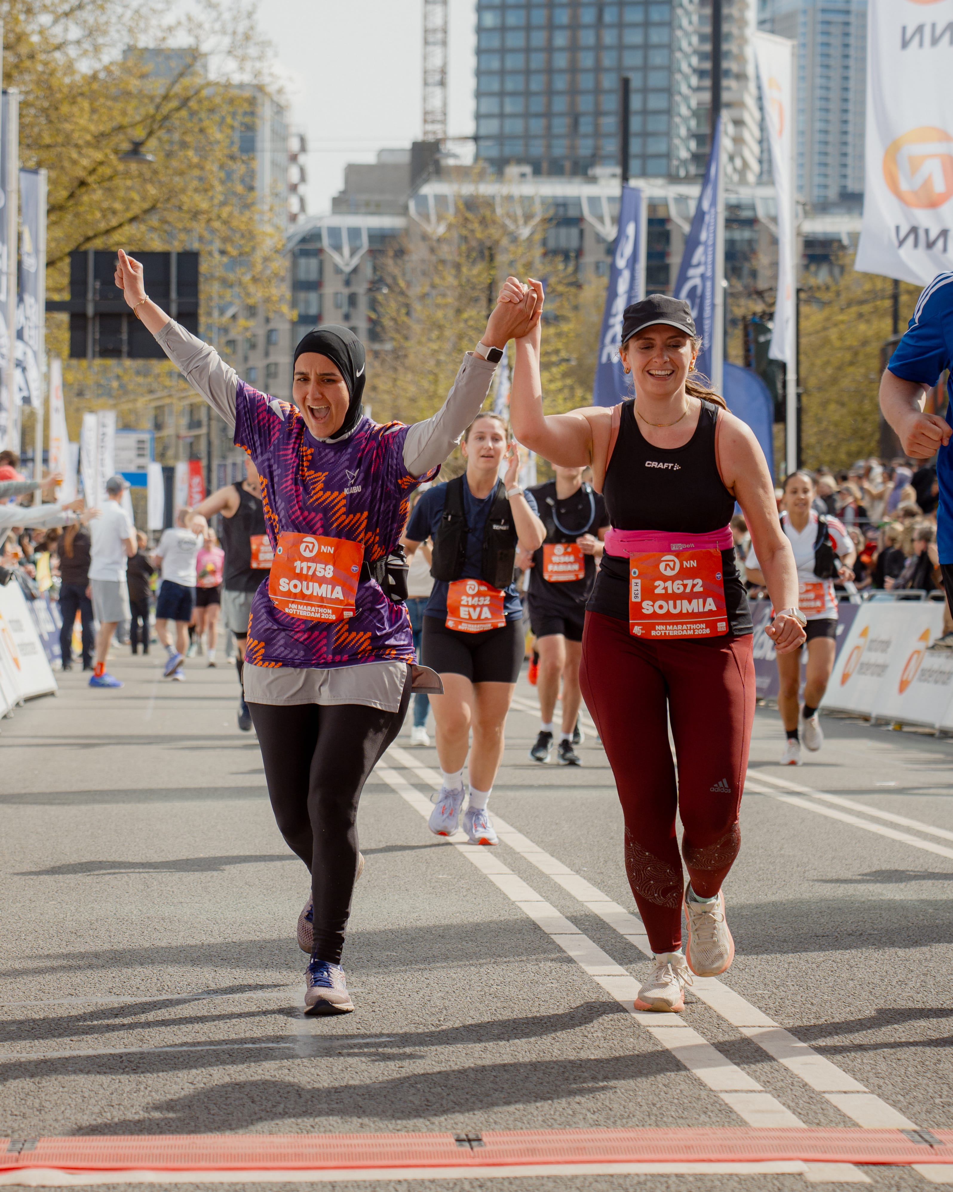 Two runners cross the marathon finish line together, smiling and holding hands, surrounded by a crowd and city buildings.
