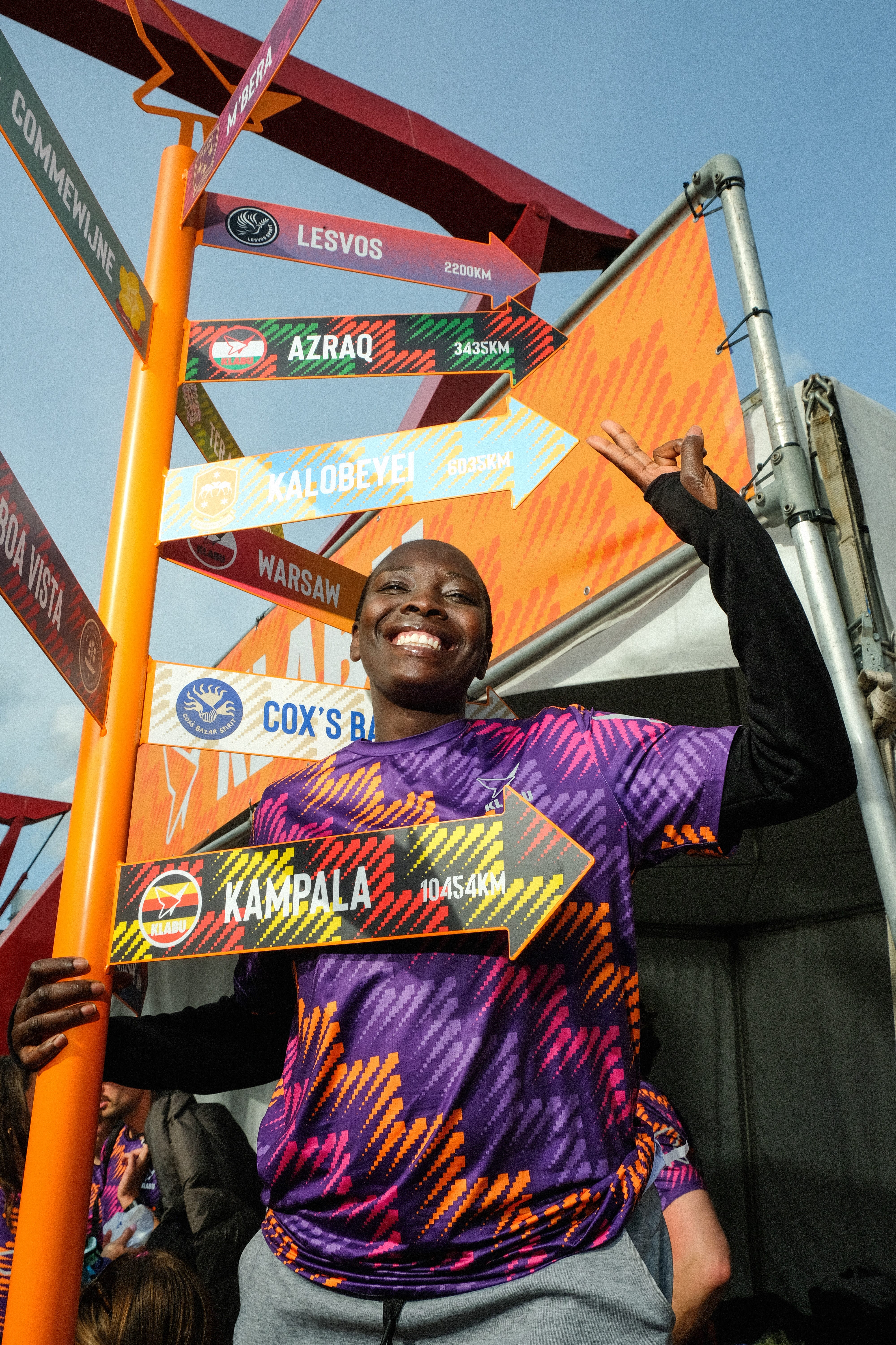 Smiling person in a colorful shirt stands next to a signpost showing distances to various locations like Kampala and Lesvos.