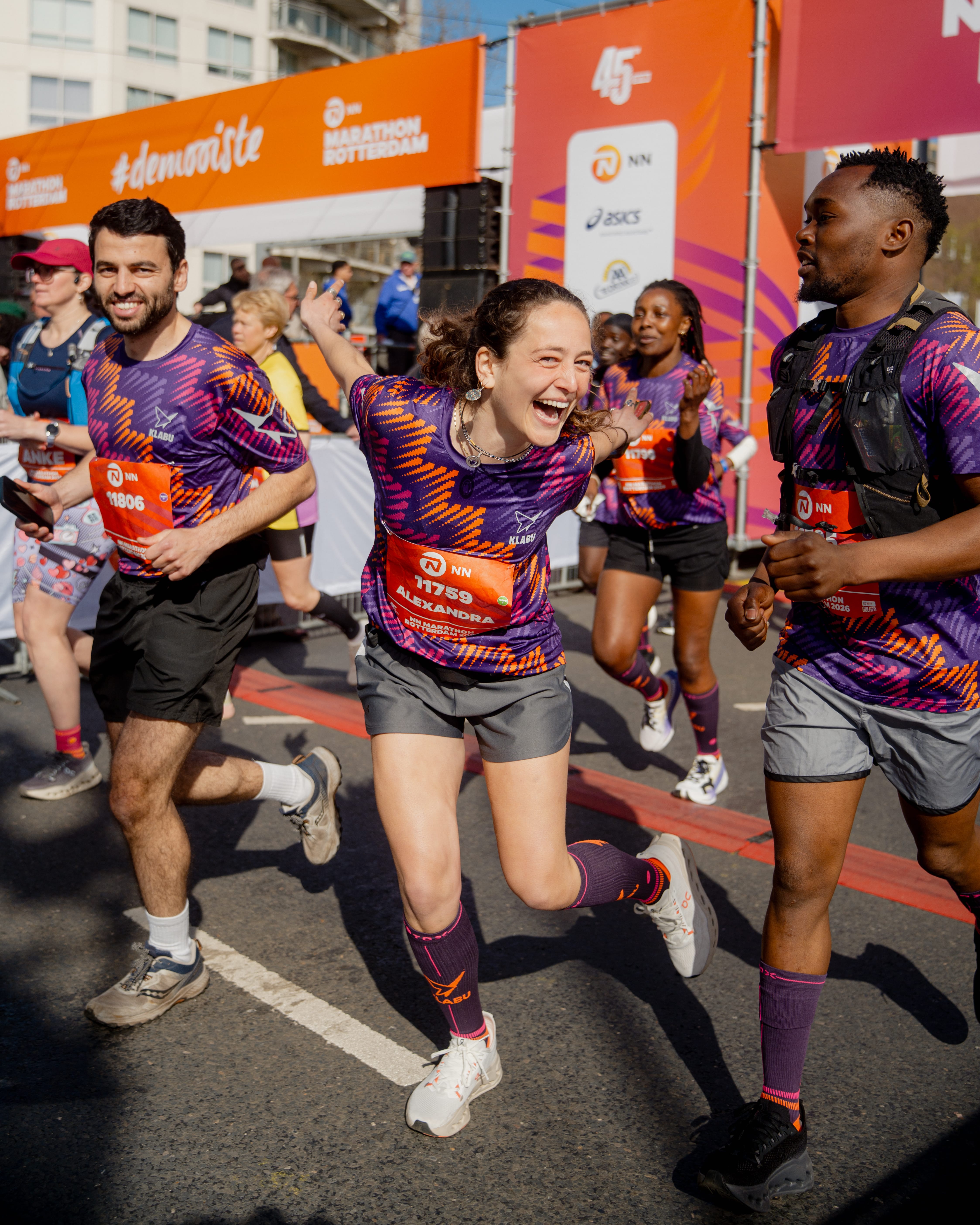 Runners in vibrant shirts joyfully sprinting during the Rotterdam Marathon, with a bright orange #demooiste banner in the background.