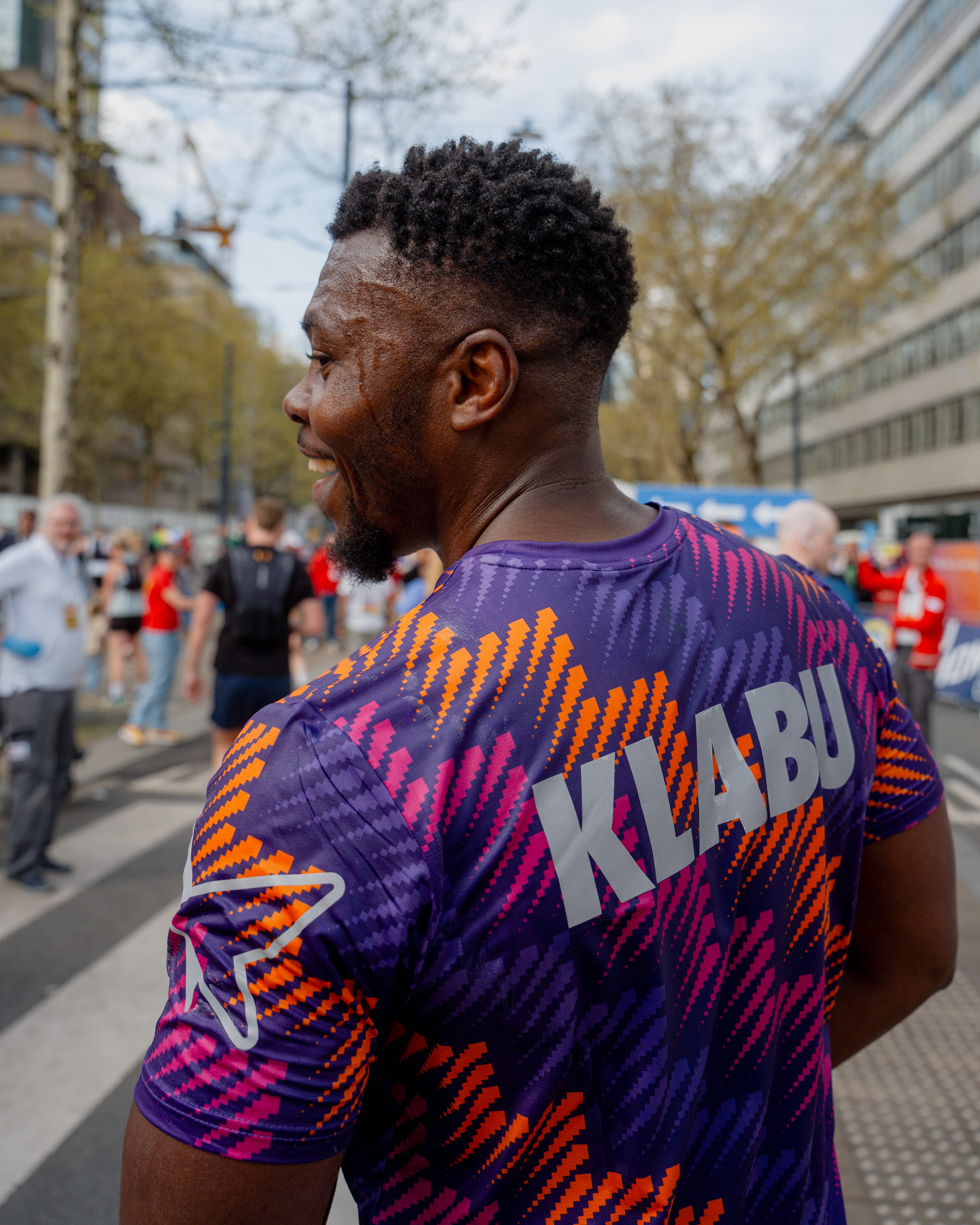 Smiling man in a colorful "KLABU" shirt stands outdoors with a blurred background of people and trees.