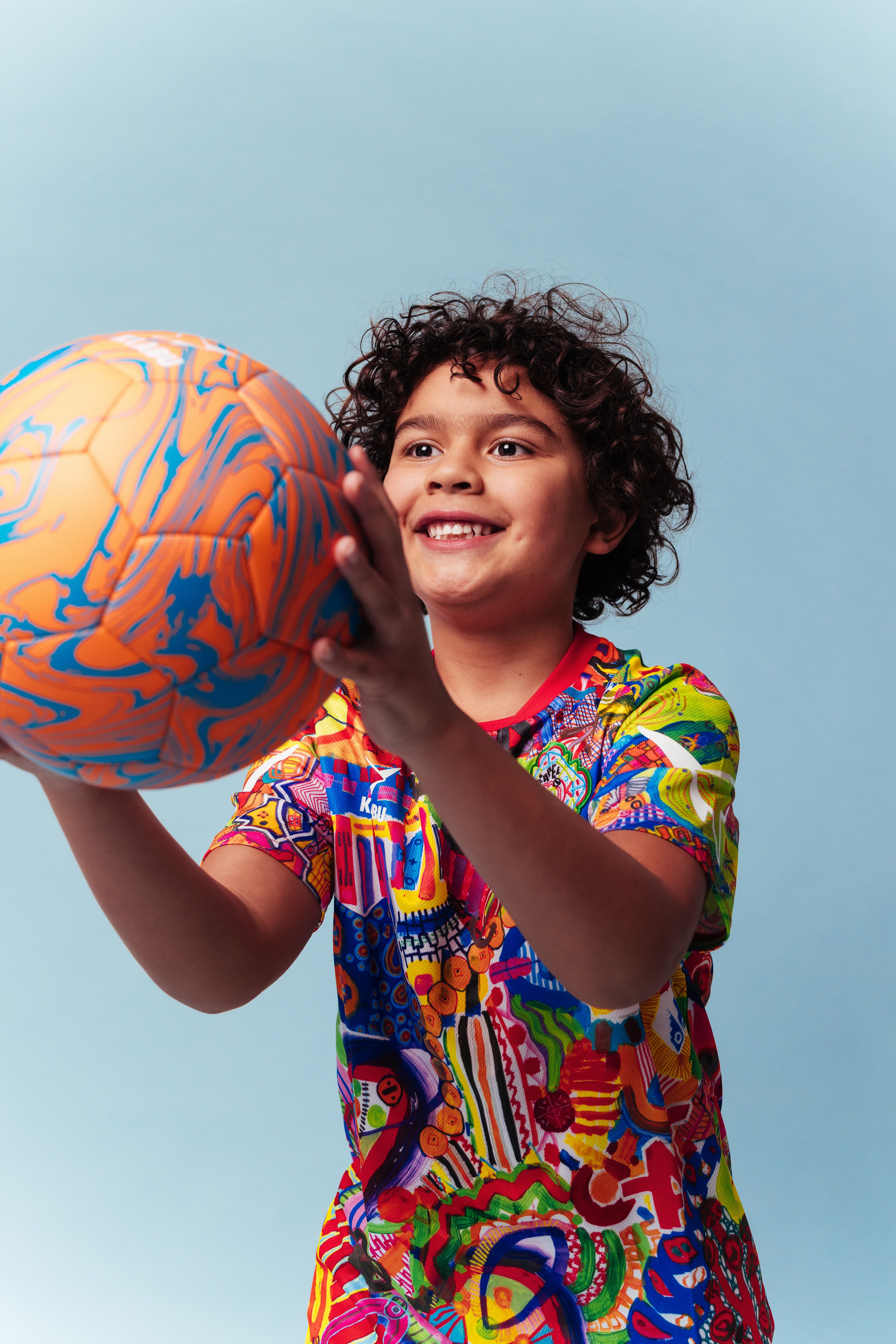 A vibrant, abstract T-shirt featuring a mix of colorful patterns, shapes, and symbols, with prominent reds, blues, yellows, and greens. Refugee sports sportswear impact