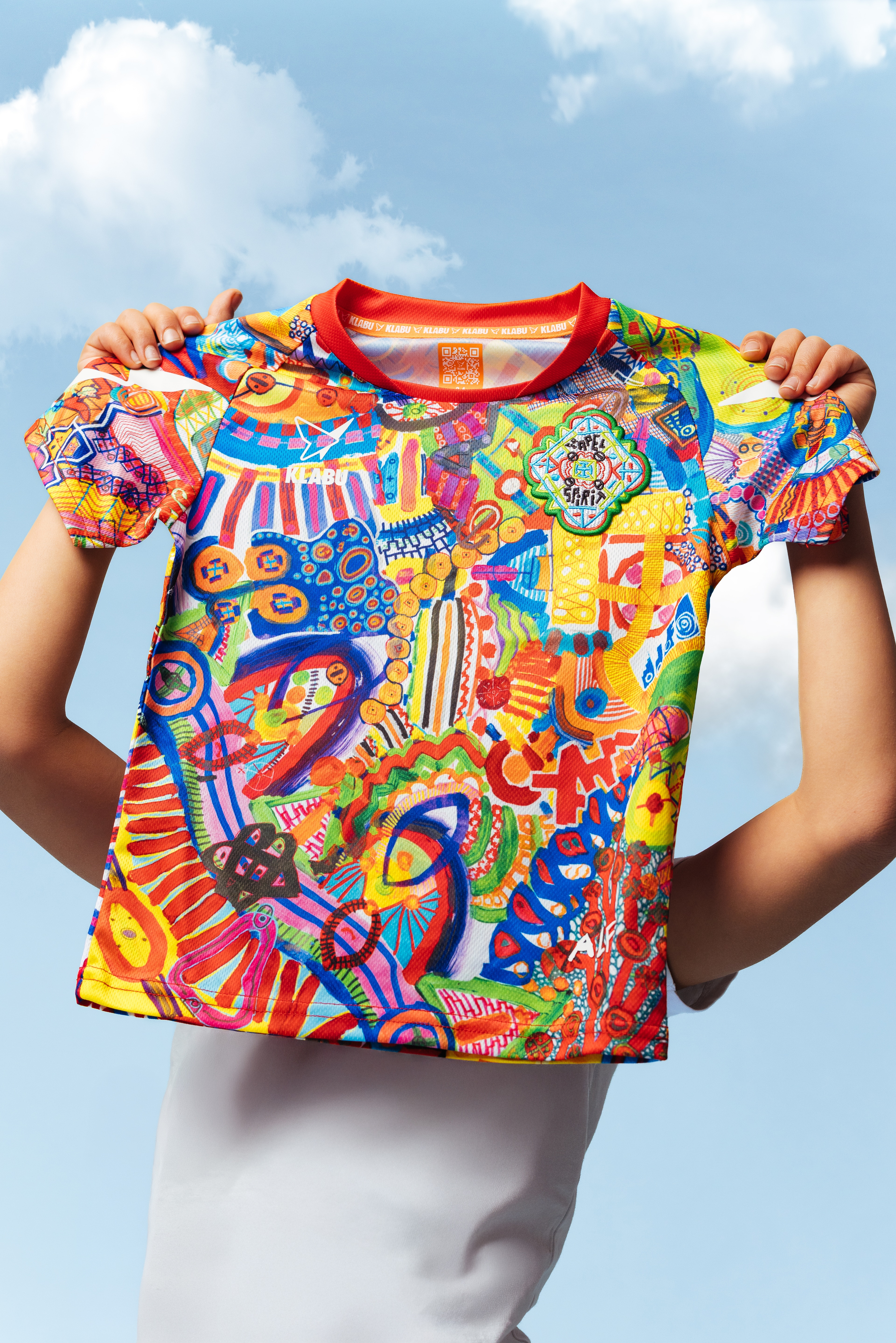 A vibrant, abstract T-shirt featuring a mix of colorful patterns, shapes, and symbols, with prominent reds, blues, yellows, and greens. Refugee sports sportswear impact