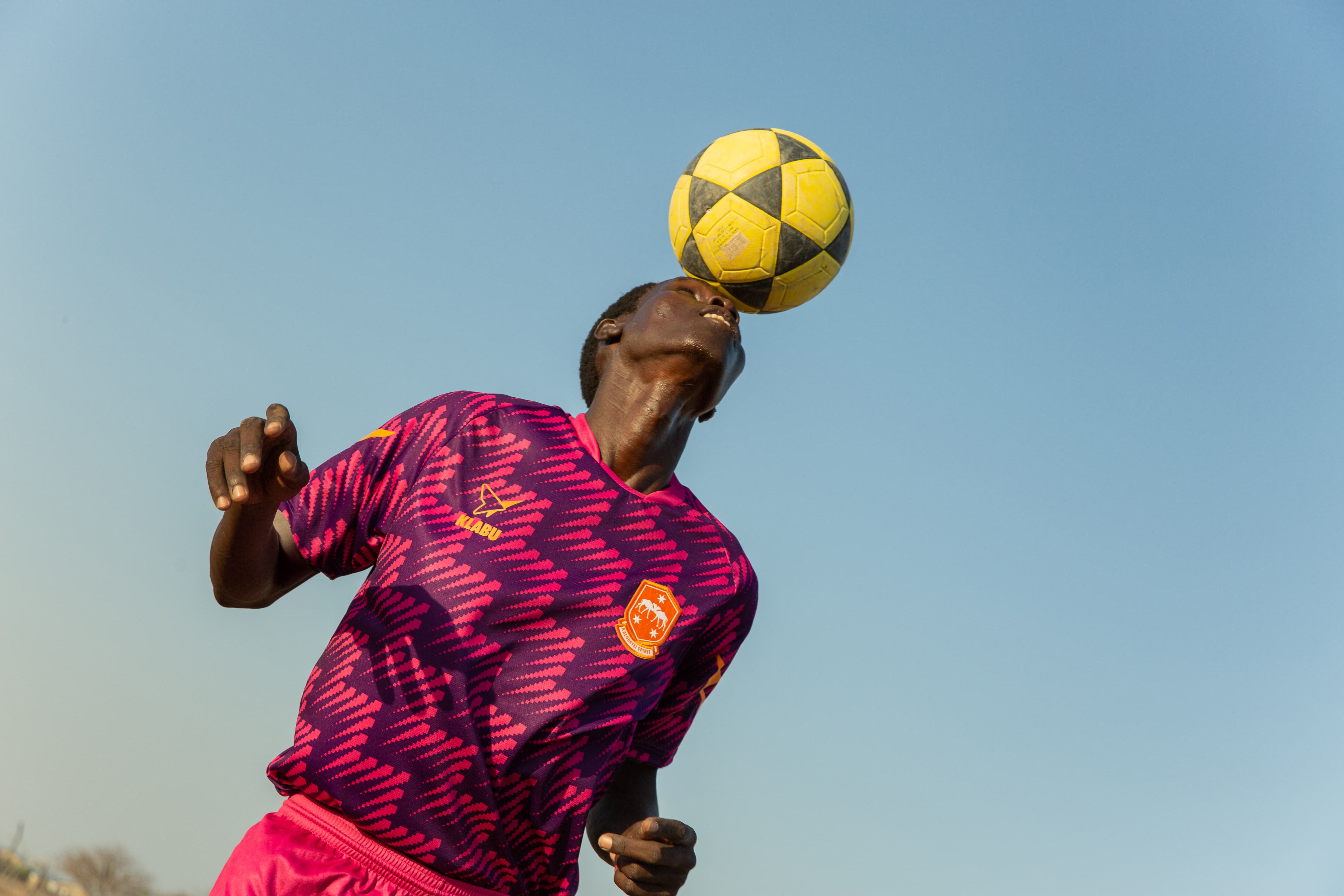 A person wearing a purple and pink jersey balances a yellow and black soccer ball on their head against a clear blue sky.