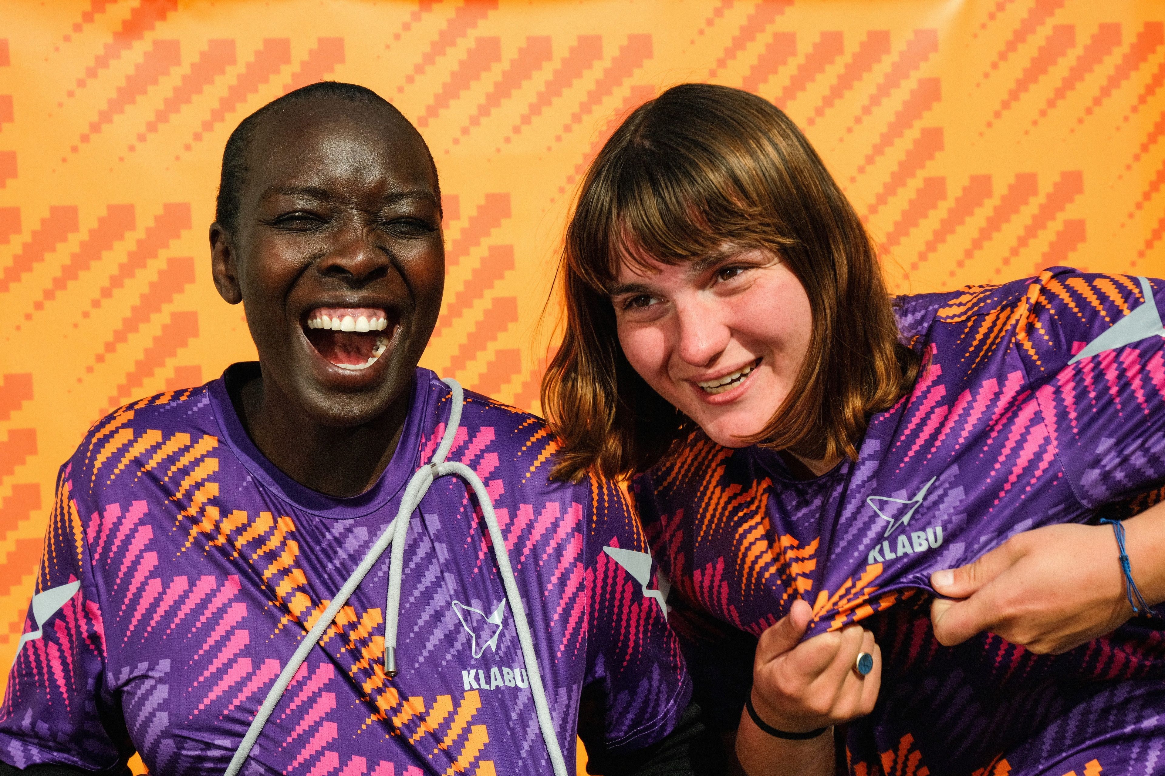 Two people joyfully smiling, wearing vibrant purple and orange "KLABU" jerseys against a patterned orange backdrop.