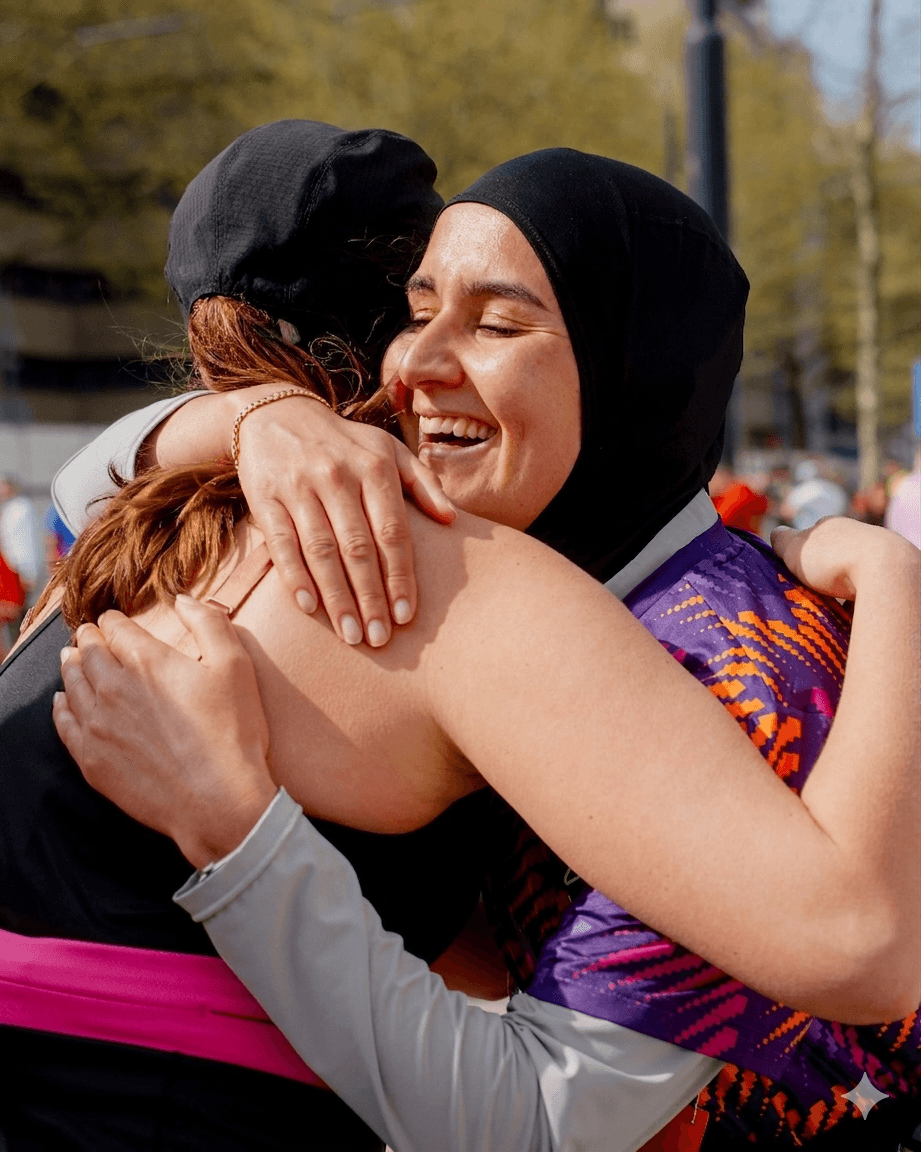 Two women embrace warmly outdoors, both smiling with joy, under a clear sky.