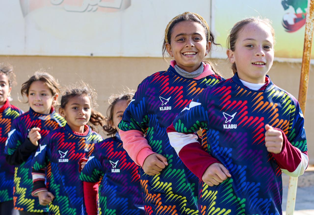 Group of young girls in colorful sports jerseys running and smiling outdoors, with a wall in the background.