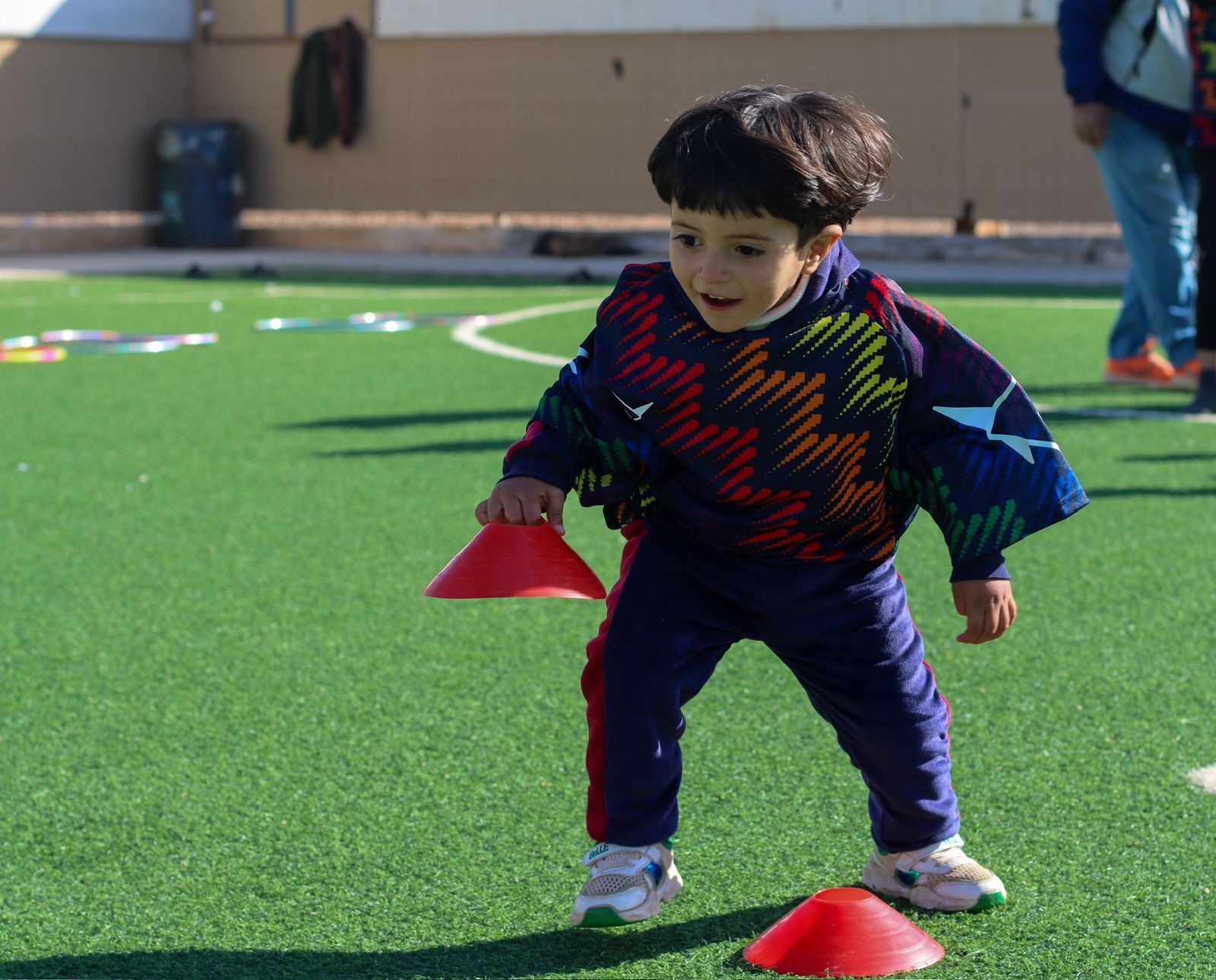 Young child in colorful clothing joyfully runs on a grass field, balancing red cones.