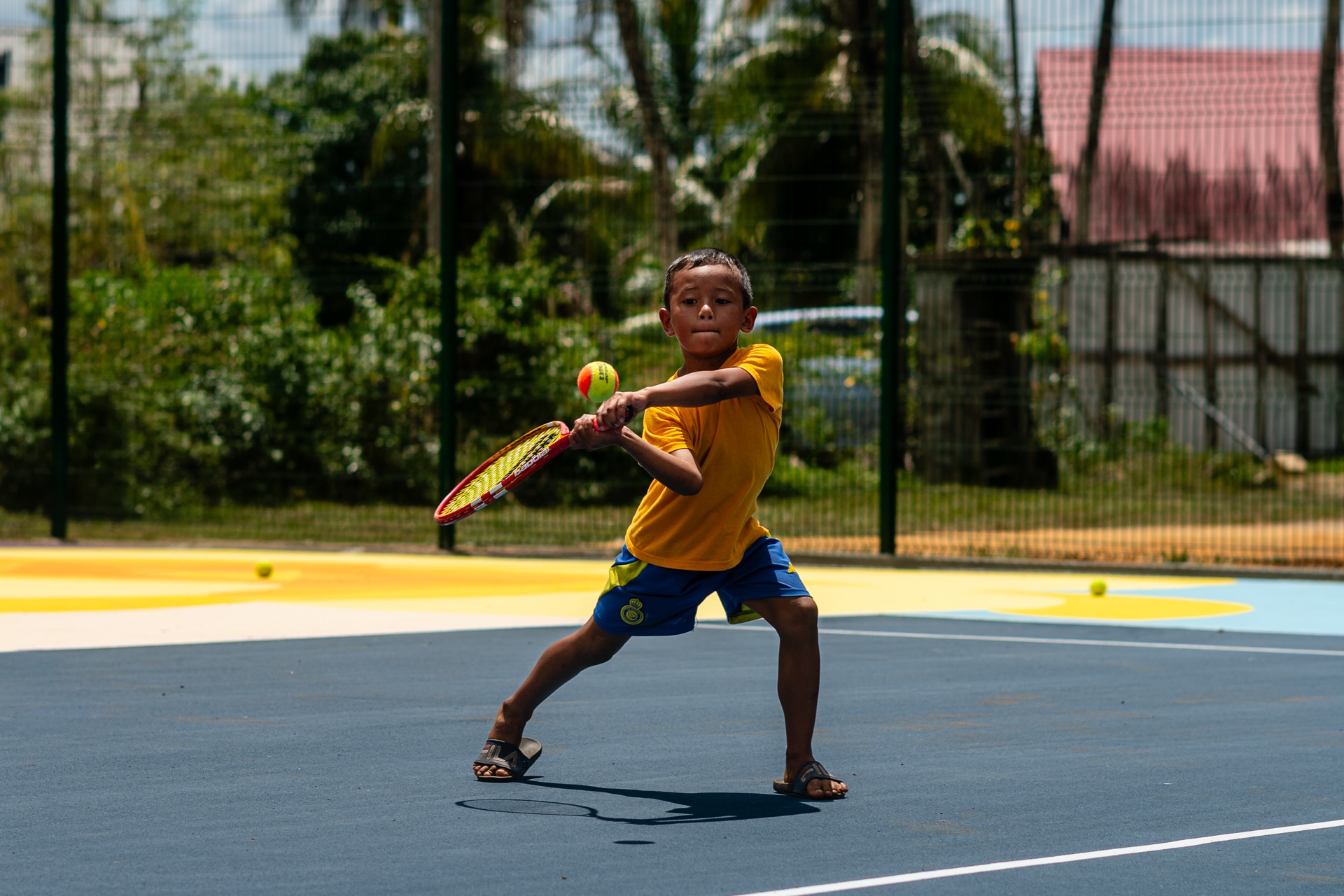 Young child in a yellow shirt and blue shorts playing tennis in Commewijne hitting a ball with a racket.