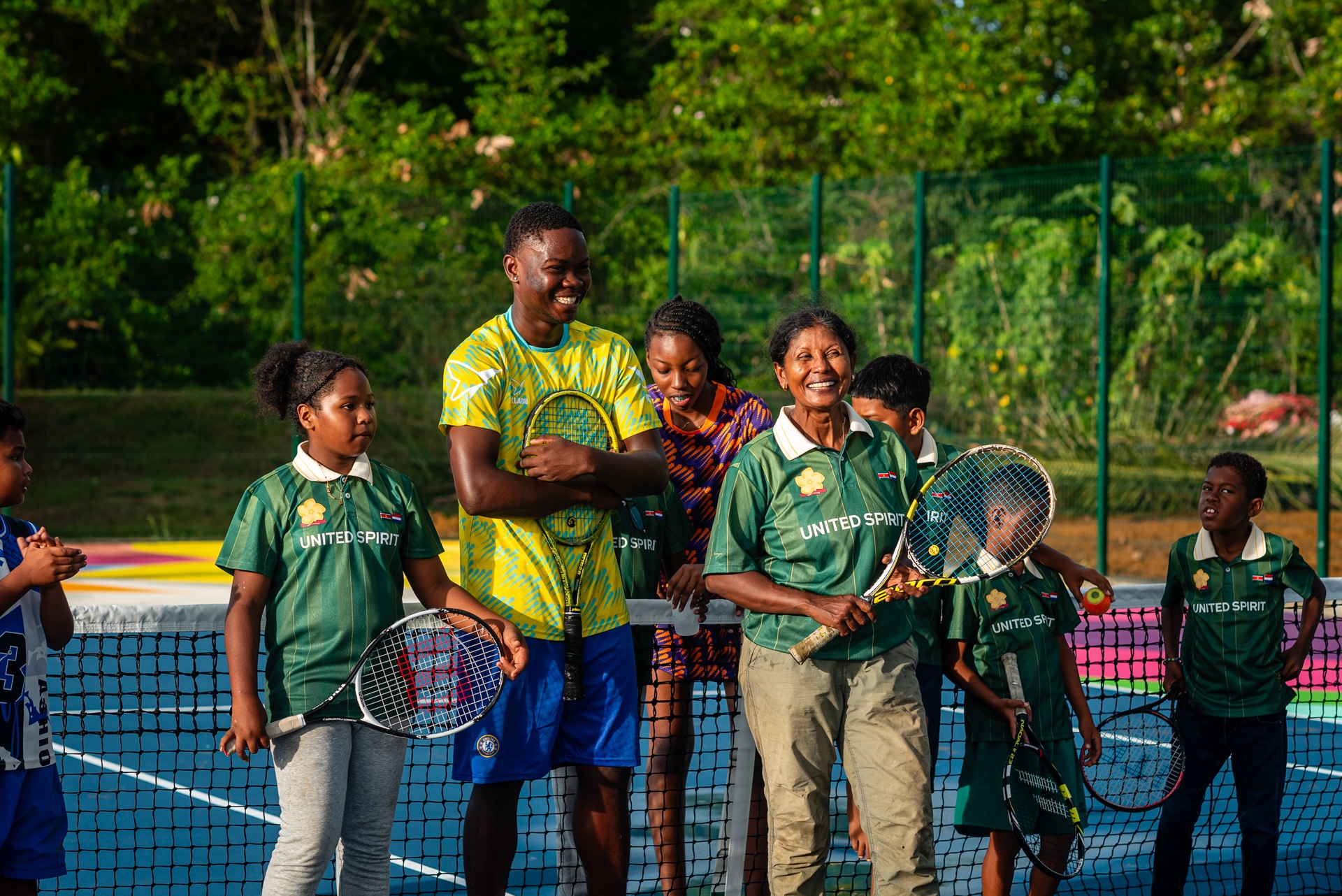 A group of people, including children and adults, stand on a tennis court holding rackets, smiling, and wearing KLABU jerseys.