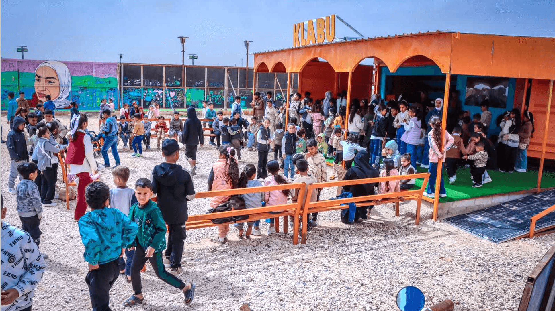 A lively outdoor gathering of children and adults near an orange pavilion, with colorful murals in the background, labeled "KLABU."