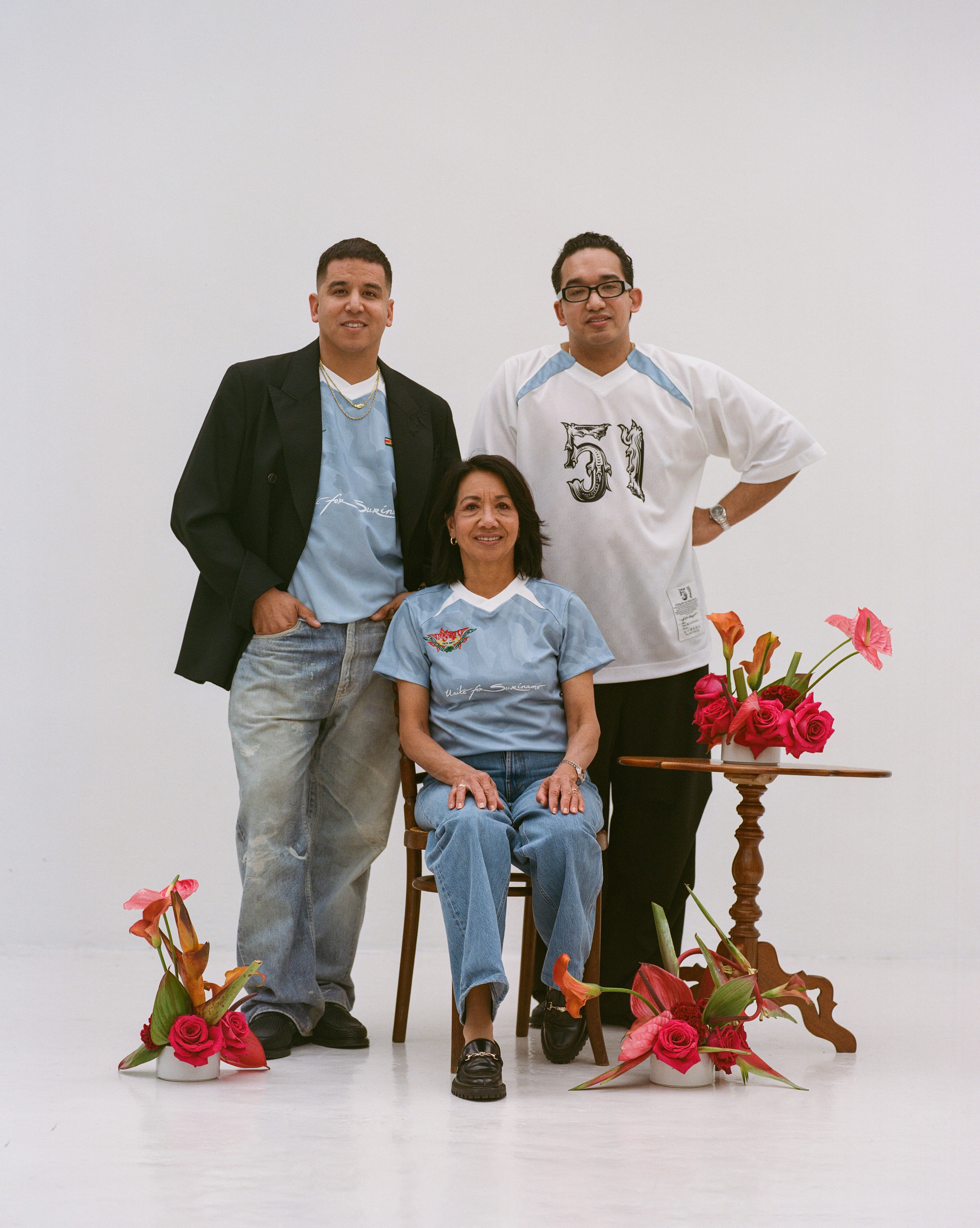 A family of 3 from Surinamese decent wearing limited edition shirts in support of Keti Koti 2026 posing next to a table with flowers surrounding them