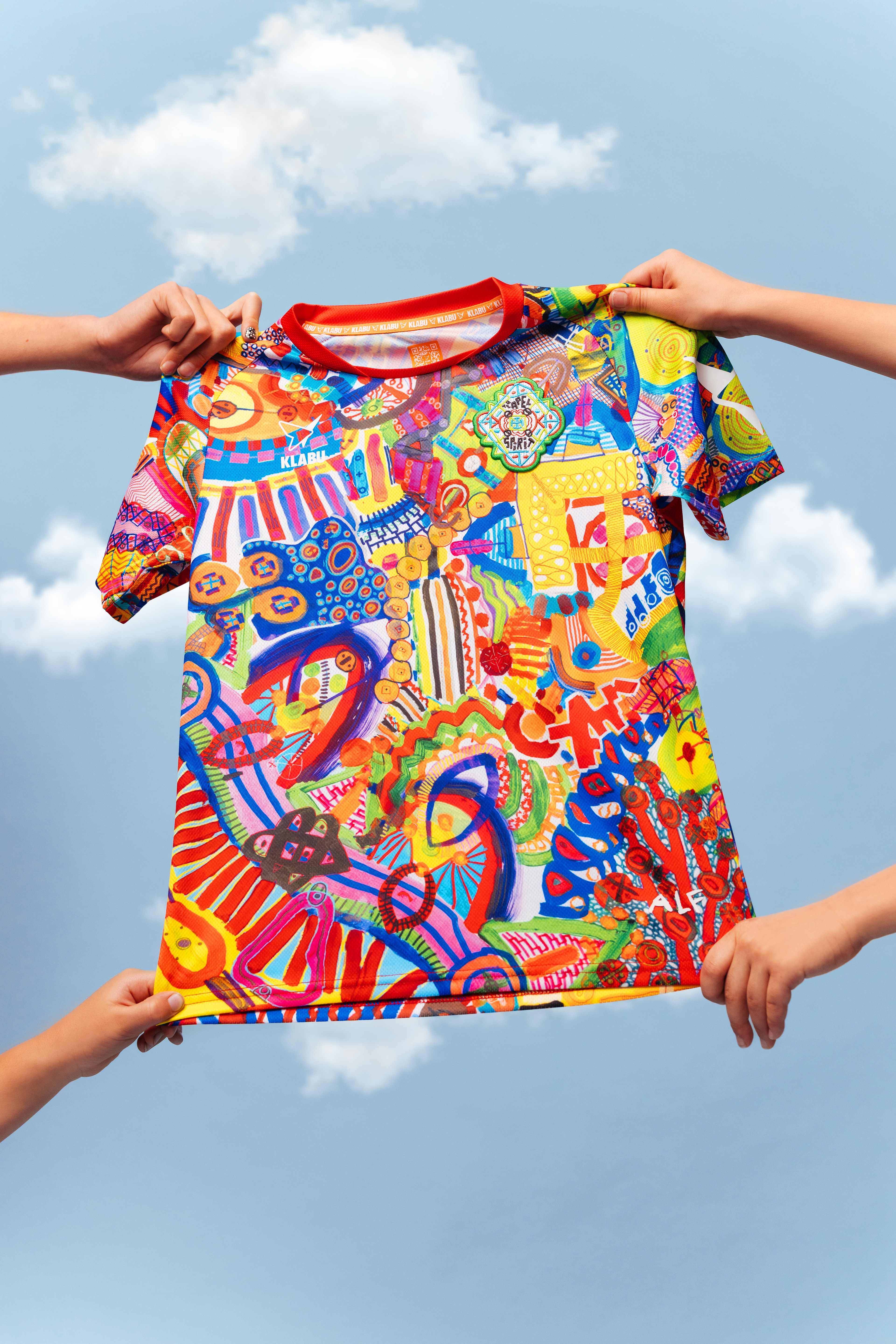 A vibrant, abstract T-shirt featuring a mix of colorful patterns, shapes, and symbols, with prominent reds, blues, yellows, and greens. Refugee sports sportswear impact