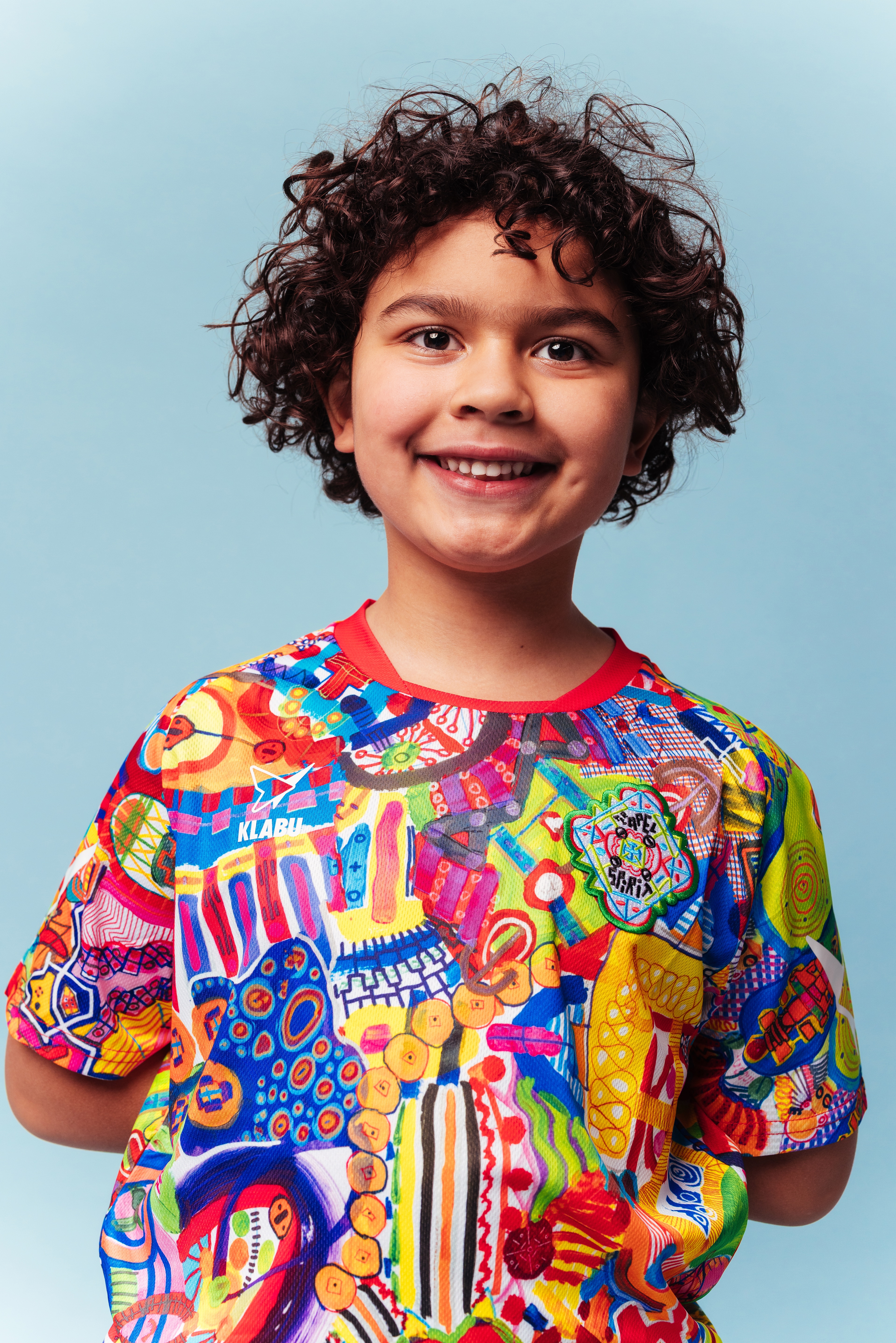 A vibrant, abstract T-shirt featuring a mix of colorful patterns, shapes, and symbols, with prominent reds, blues, yellows, and greens. Refugee sports sportswear impact